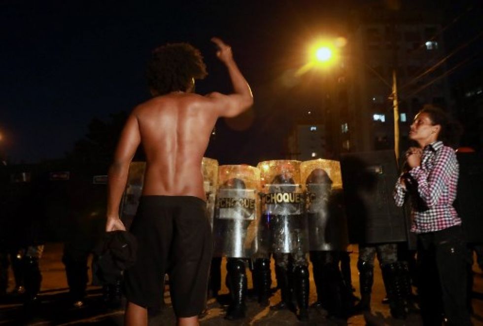 Un hombre negro se enfrenta a la policía antidisturbios mientras una mujer negra reza durante una protesta.
