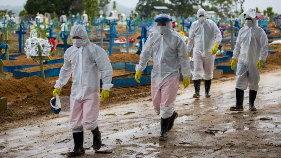 Trabajadores vestidos con trajes protectores caminan junto a las tumbas de las víctimas del covid-19 en un cementerio brasileño.