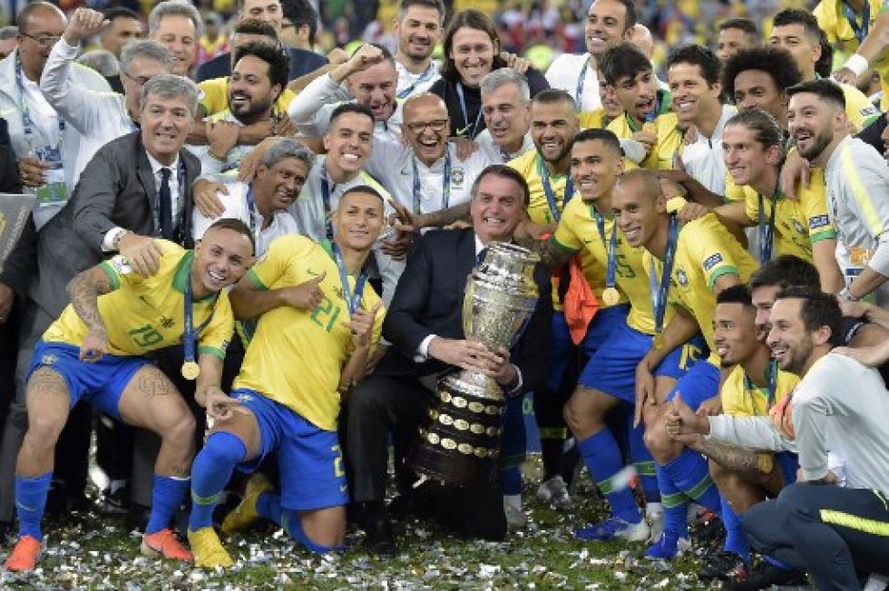 El presidente brasileño Jair Bolsonaro sostiene el trofeo de la Copa América mientras miembros de la selección brasileña celebran tras ganar el título al derrotar a Perú en 2019.